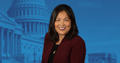 Julie Su in the foreground with U.S. government buildings in the background with a blue overlay